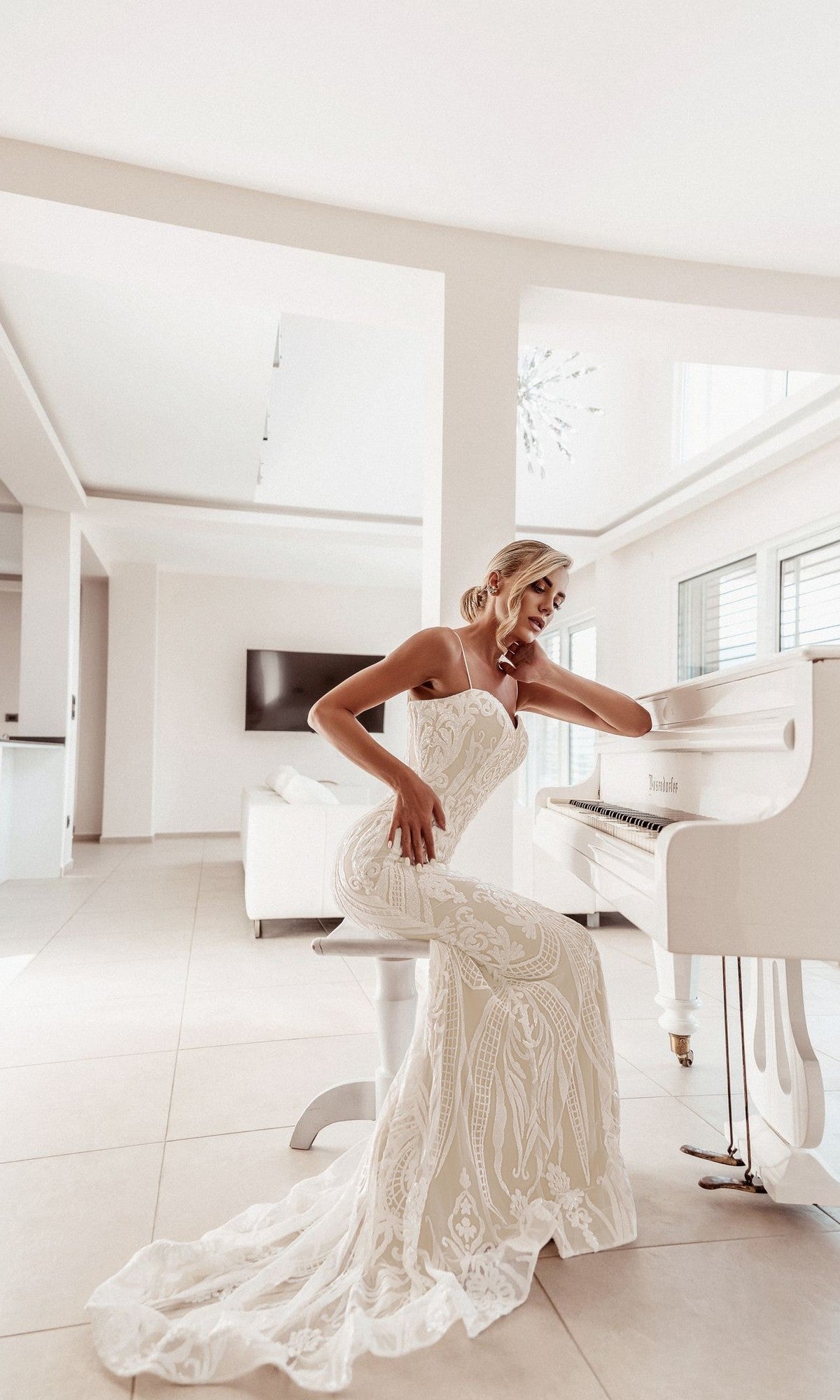 A woman in the Tina Holly BA999 White Lace Bodycon Bridal Gown sits on a white stool next to a white grand piano in a bright, modern living room.